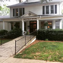 Exterior of the Wellness in downtown Winston-Salem. Collective on 5th Street. A two story white house with a wide porch bordered by shrubs. A short portico has the words "Troutman House" in black lettering.  A concrete accessibily ramp extends from the porch to the sidewalk. The ramp is flanked by a smal grass lawn.